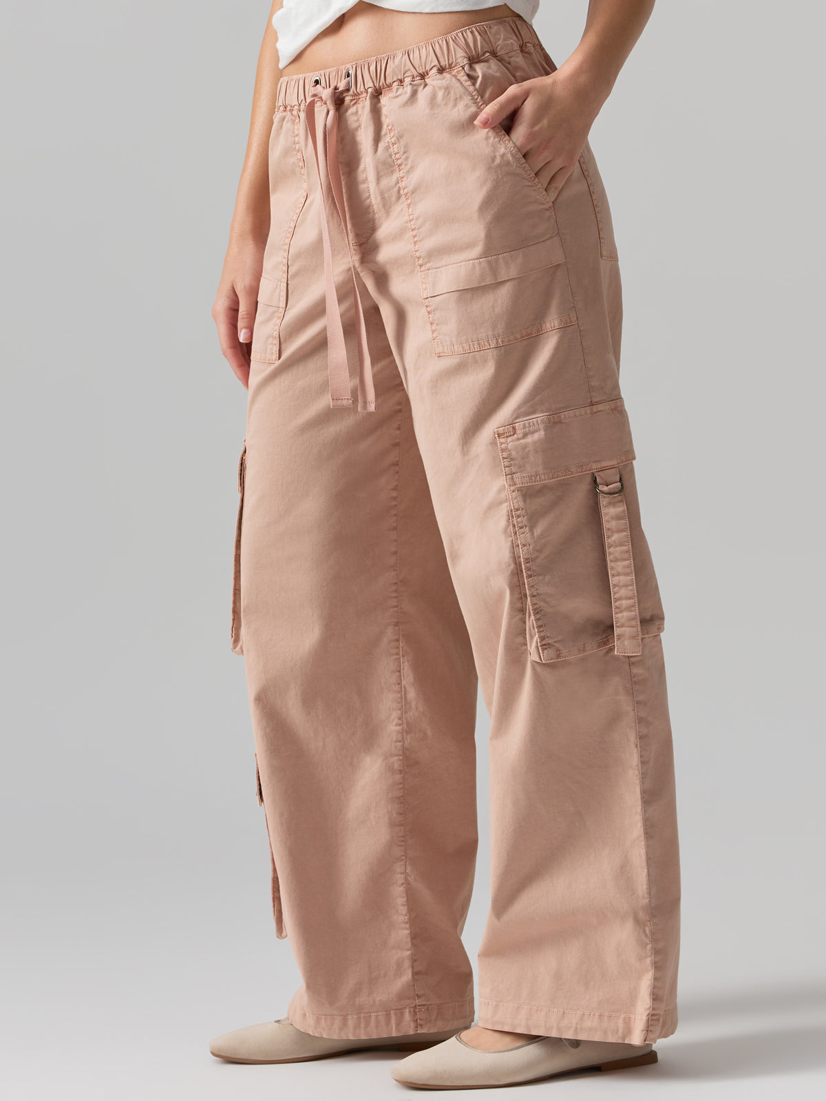 A person wearing Sanctuary Clothing's Cargo Parachute Pant Bare Nude, which are loose-fitting with multiple large leg pockets and an elastic waistband with a drawstring. They are posing with one hand in a pocket, wearing a white top and beige shoes against a plain gray background.