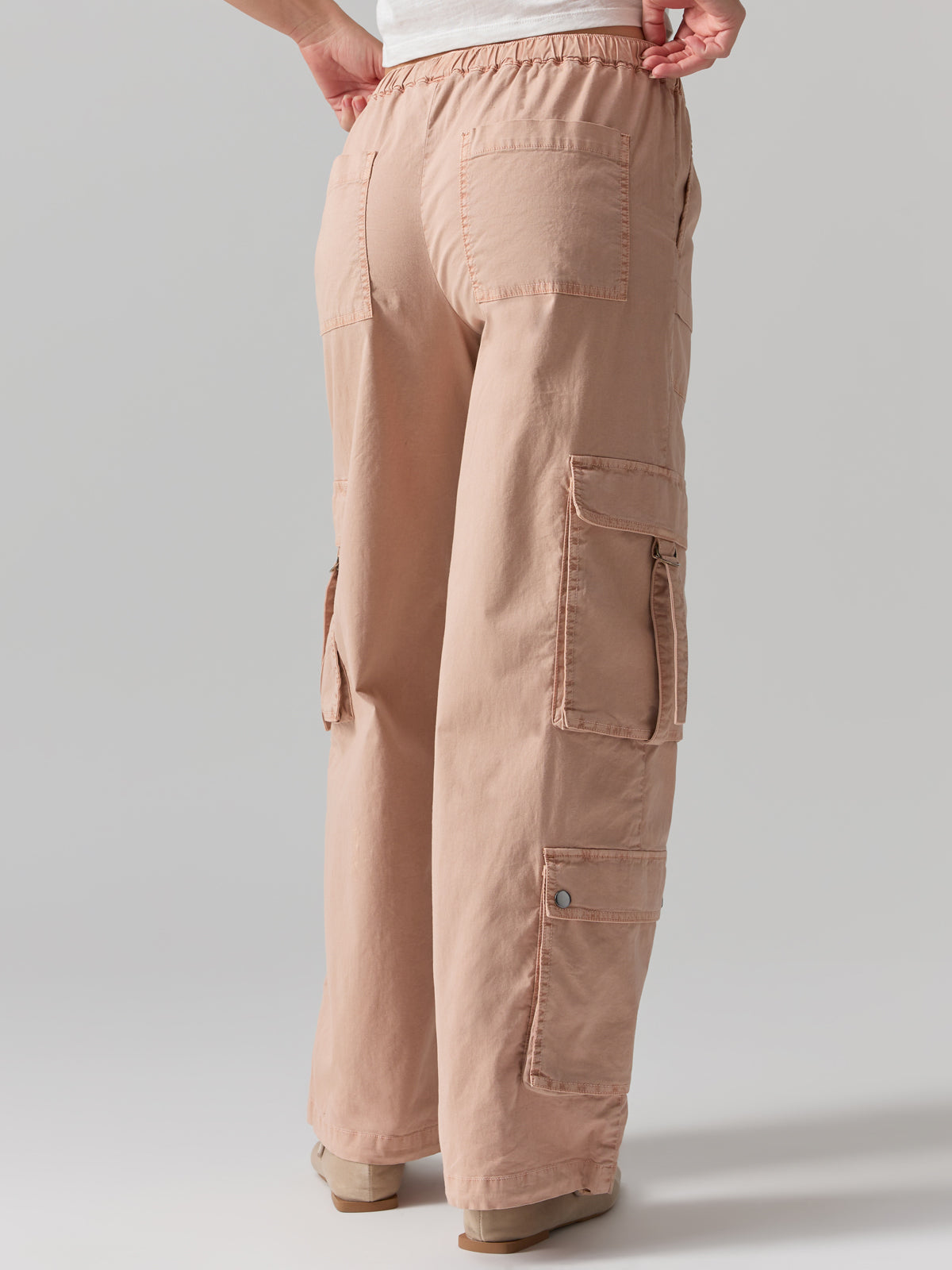 A person is standing with their back to the camera, wearing the Cargo Parachute Pant Bare Nude from Sanctuary Clothing, featuring multiple functional pockets and a white waistband. The photo highlights the design of these light brown pants, including their loose fit and practical pockets, against a plain, light-colored background.