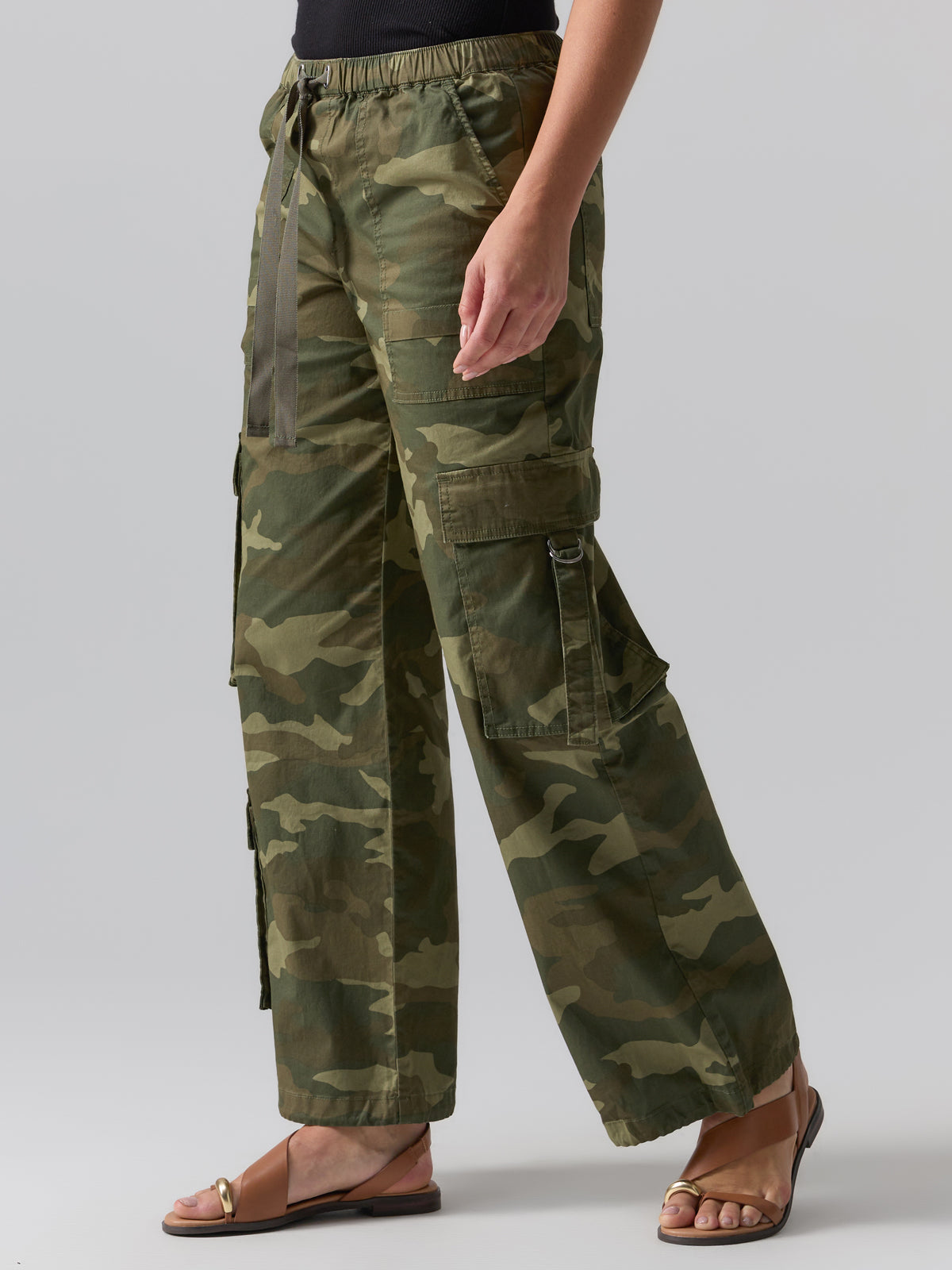 A person is dressed in Sanctuary Clothing's Cargo Parachute Pant Renew Camo, a green camouflage cargo pant with a loose fit and multiple side pockets. They are also wearing brown sandals and a black top. The background is plain gray, showing only the lower half of their body.