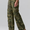 A person is dressed in Sanctuary Clothing's Cargo Parachute Pant Renew Camo, a green camouflage cargo pant with a loose fit and multiple side pockets. They are also wearing brown sandals and a black top. The background is plain gray, showing only the lower half of their body.