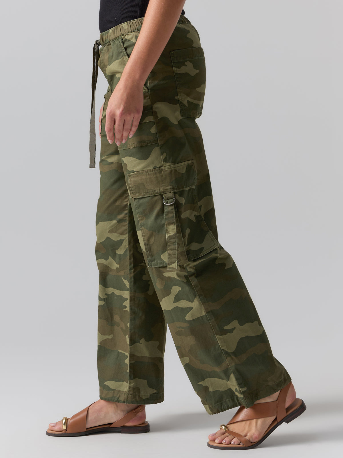 The photo features a person wearing the Cargo Parachute Pant Renew Camo by Sanctuary Clothing and brown sandals. Captured from a side view, the image shows the lower half of their body adorned in loose-fitting, green camouflage cargo pants with multiple pockets. A black top is partly visible in the shot against a plain gray background.