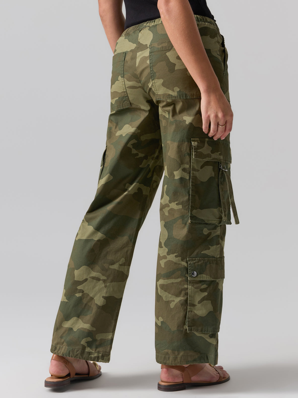 A person wearing Sanctuary Clothing's Cargo Parachute Pant Renew Camo stands facing away from the camera. The green camouflage pants feature multiple pockets, including side cargo pockets with flaps and buttons. The individual is also sporting a black top and brown sandals against a plain gray background.