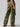 A person wearing Sanctuary Clothing's Cargo Parachute Pant Renew Camo stands facing away from the camera. The green camouflage pants feature multiple pockets, including side cargo pockets with flaps and buttons. The individual is also sporting a black top and brown sandals against a plain gray background.