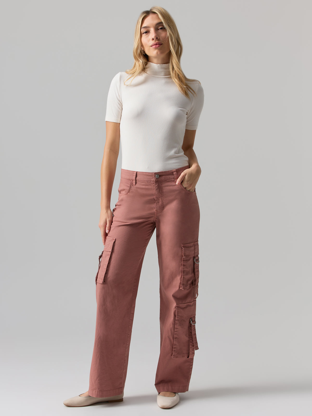 A person with long blonde hair stands against a plain background, wearing a fitted white short-sleeve turtleneck and the Y2K Strappy Pant Ash Rose by Sanctuary Clothing. They look at the camera with a relaxed expression, their right hand in the pocket of the high-waisted pink cargo pants with side pockets.