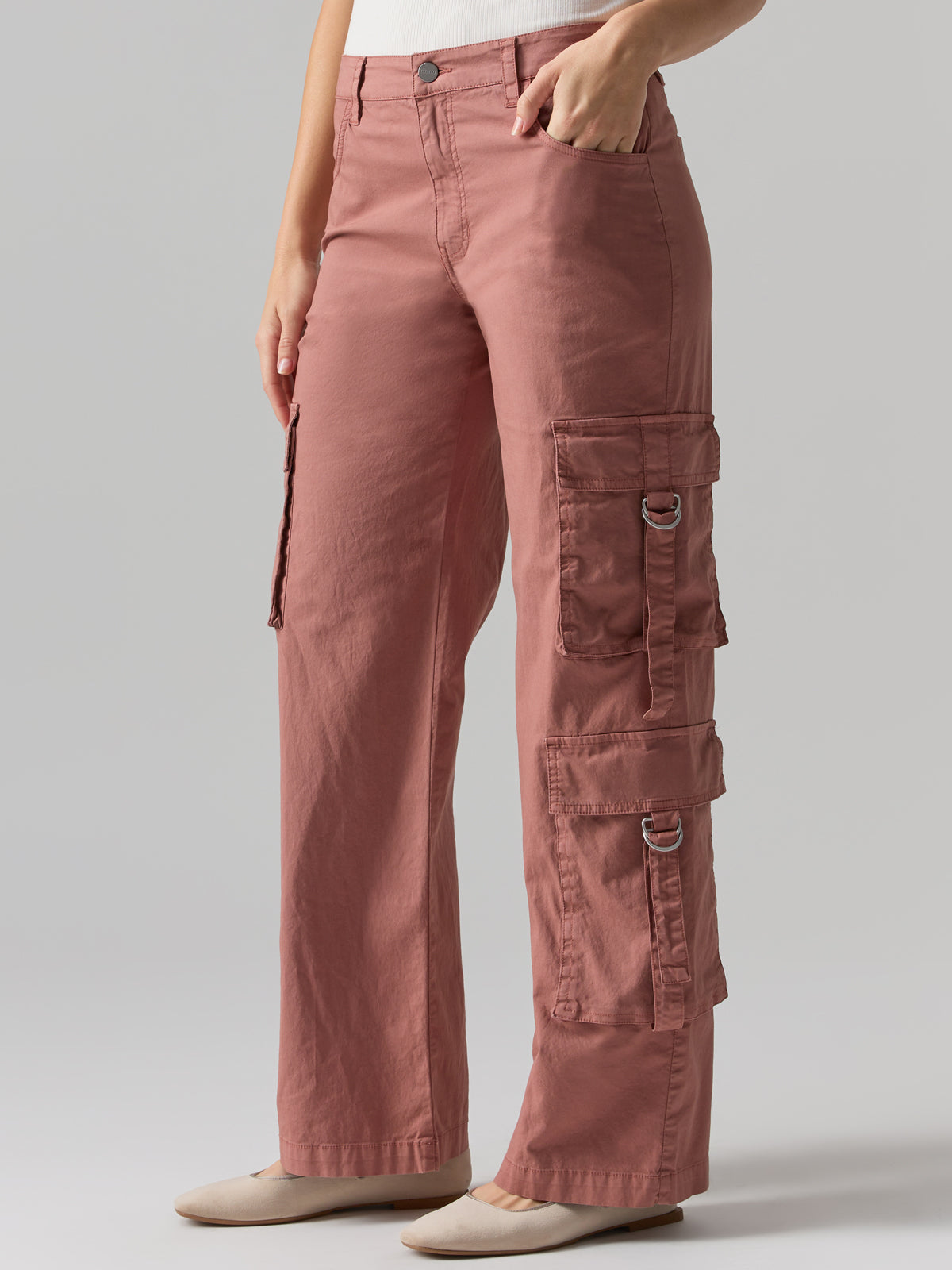 A person wearing the Y2K Strappy Pant Ash Rose by Sanctuary Clothing, featuring multiple pockets and metal rings on the sides. The pants are paired with light-colored footwear. The person is standing against a plain, light gray background, with only the lower half of their body visible.