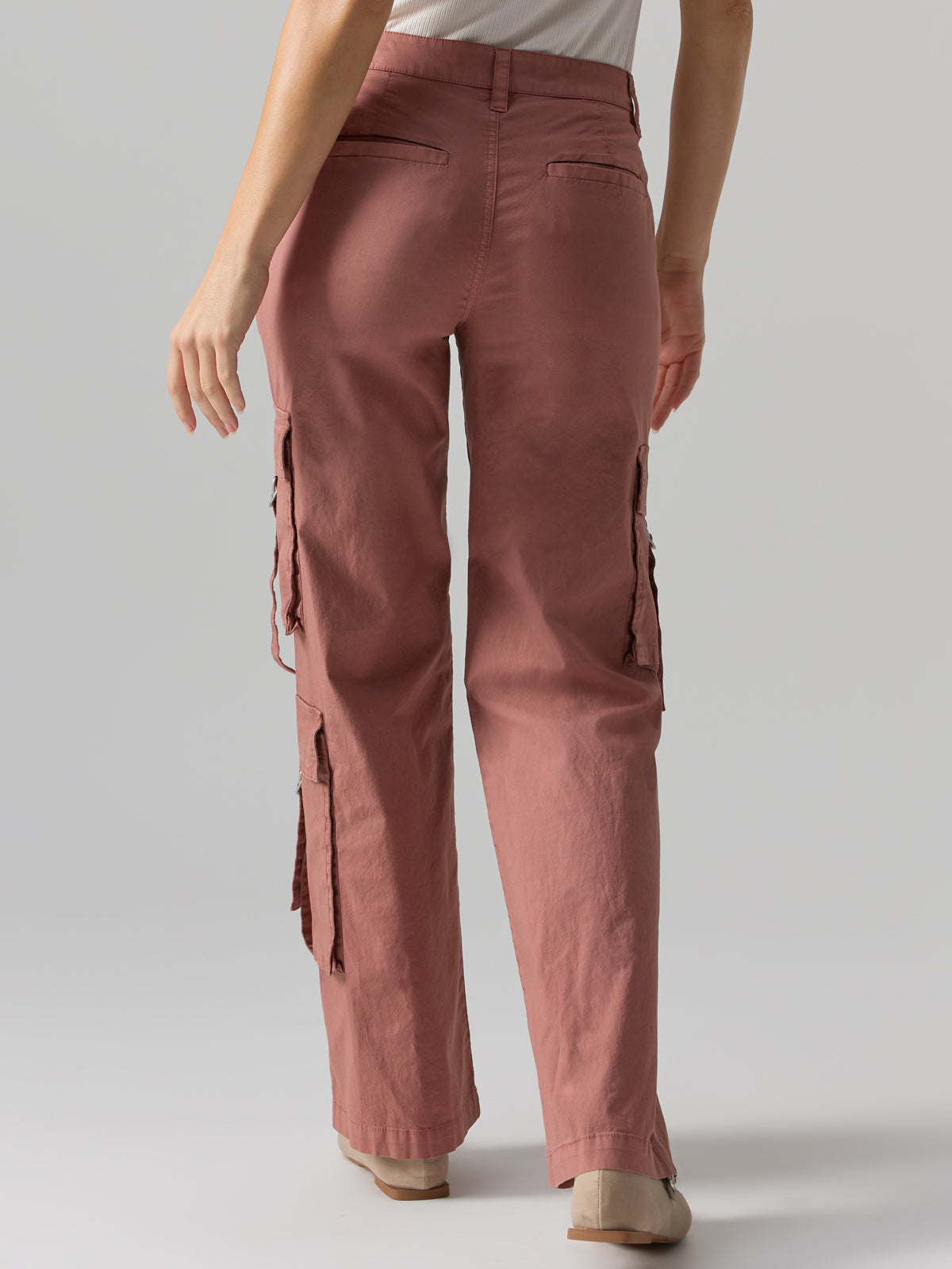 A person wearing the Sanctuary Clothing Y2K Strappy Pant Ash Rose, featuring high-waisted, rose-colored fabric and side cargo pockets, is showing the back view. The pants have a slightly loose fit and are paired with a white top and beige shoes.