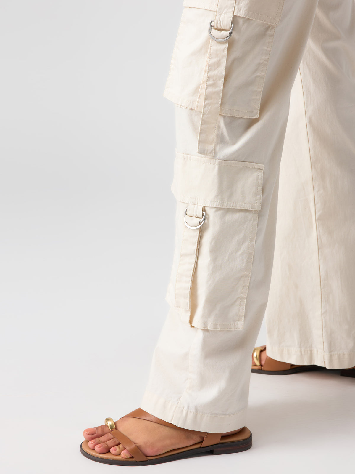 Close-up of a person wearing Sanctuary Clothing's Y2K Strappy Cargo Pant Eco Natural with multiple pockets and metal D-rings, paired with brown sandals featuring a metal accent on the toe strap, standing against a plain background.