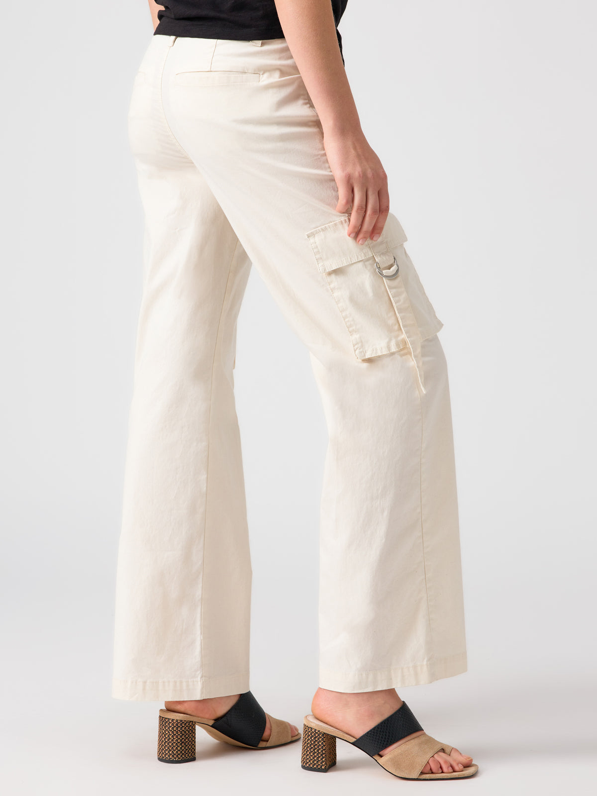 A person is wearing the Sanctuary Clothing Y2K Strappy Cargo Pant Eco Natural, which are cream-colored wide-leg cargo pants featuring a large pocket on the left side, paired with black block-heeled sandals. The upper body is out of frame, and the background is plain light gray.