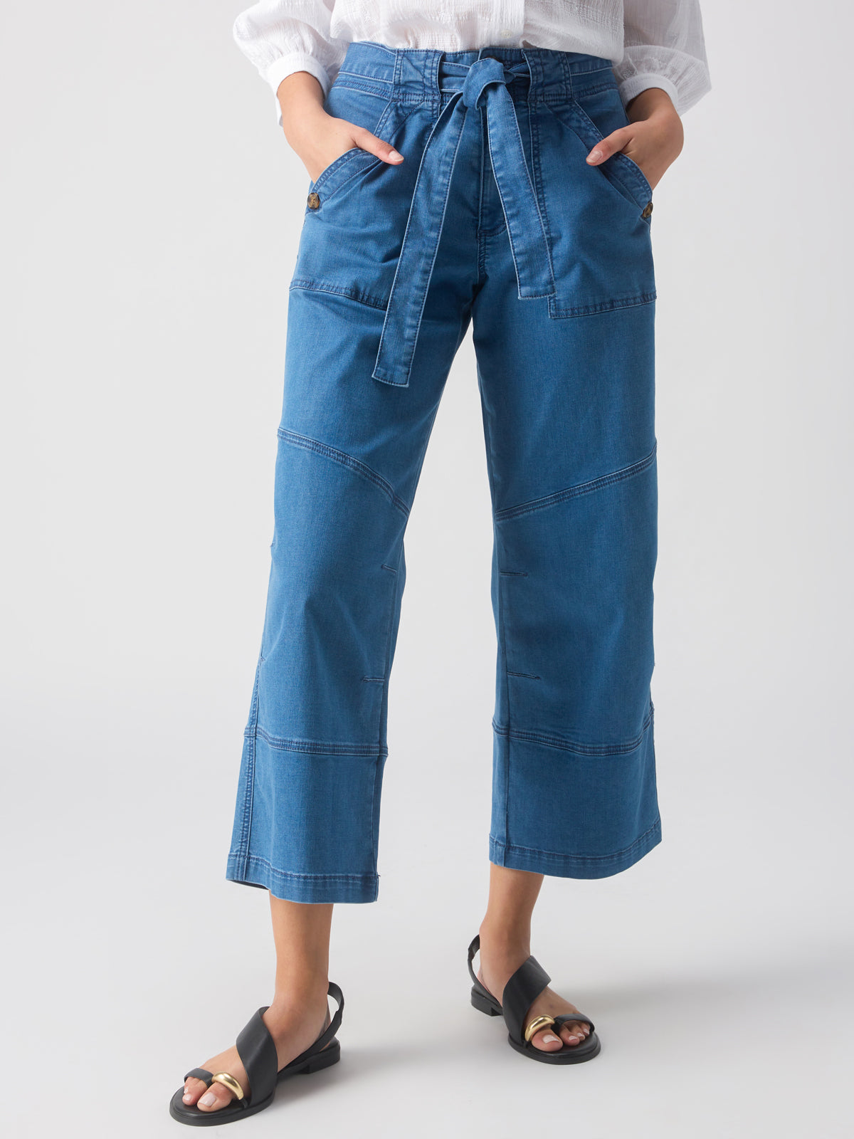 A person wearing the Reissue 90S Sash Spring Valley jeans from Sanctuary Clothing, which are blue high-waisted wide-leg jeans with visible stitching details and a belted waist, is standing with hands in pockets. They are also wearing black open-toe sandals and a white top. The background is plain and light-colored.