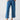 A person wearing the Reissue 90S Sash Spring Valley jeans from Sanctuary Clothing, which are blue high-waisted wide-leg jeans with visible stitching details and a belted waist, is standing with hands in pockets. They are also wearing black open-toe sandals and a white top. The background is plain and light-colored.