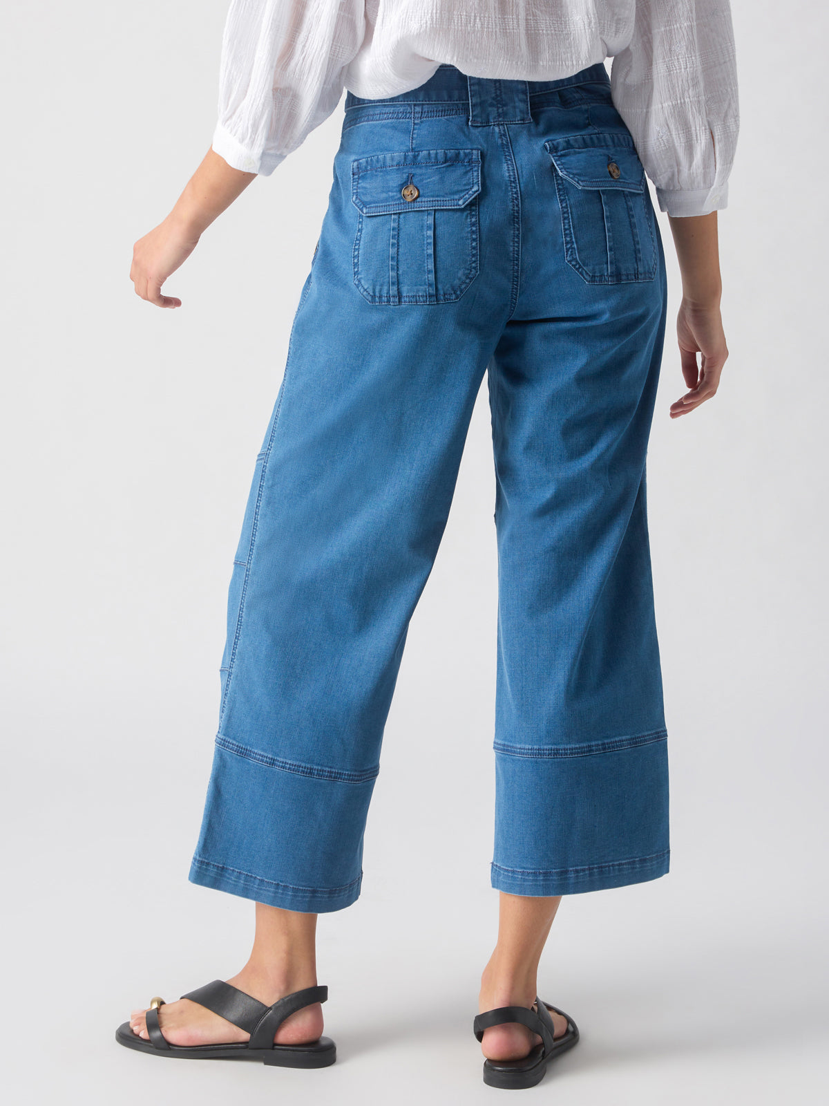 A person wearing the Sanctuary Clothing Reissue 90S Sash Spring Valley wide-leg, high-waisted blue jeans with buttoned back pockets and a white blouse. They're standing on a light gray background with their back to the camera and their left arm slightly bent. They're also wearing black sandals.