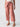 A person is seen from the waist down wearing Sanctuary Clothing's Reissue 90S Sash Ash Rose, high-waisted, wide-leg pink trousers with large front pockets and a tied belt. They are also wearing a white top and brown sandals against a plain white background.