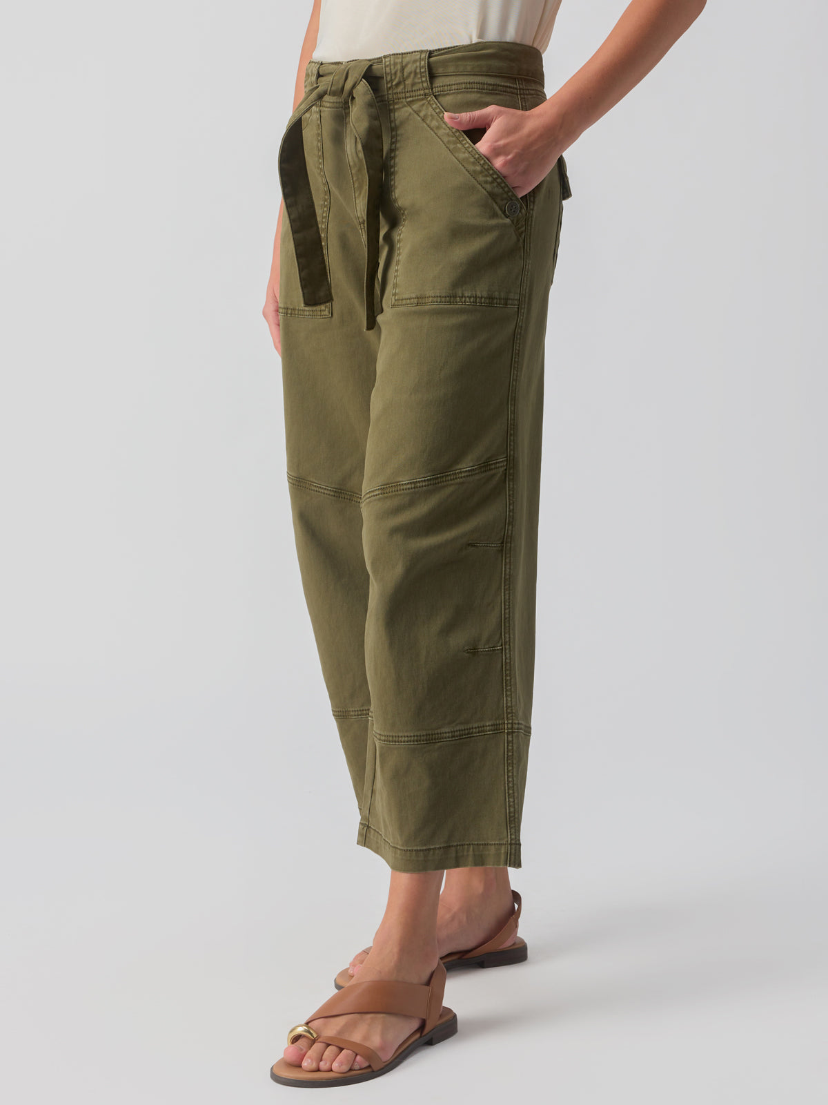 A person is wearing the Reissue 90S Sash Burnt Olive cargo pants from Sanctuary Clothing, featuring large front pockets and a fabric tie belt. They are also sporting a light-colored top and tan sandals. The photo captures them from the waist down against a plain background.