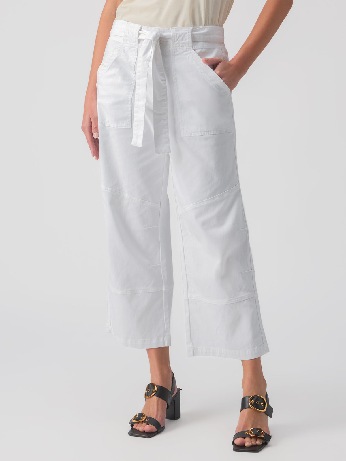 A person wearing the Reissue 90S Sash White pants by Sanctuary Clothing, which are white, wide-legged, cropped with a tied waist, and sandals with black straps and gold buckles. The pants feature large front pockets and seam details.