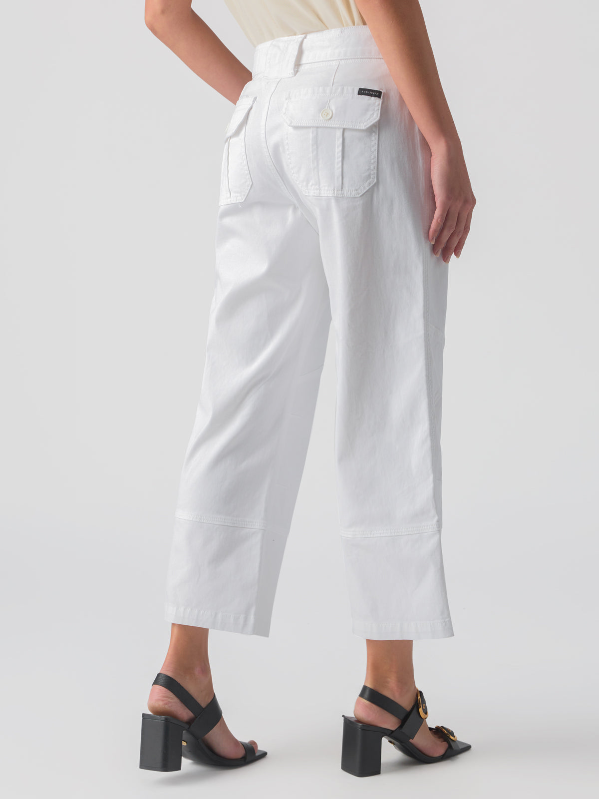 A person is wearing the Reissue 90S Sash White high-waisted, wide-leg pants from Sanctuary Clothing, featuring side and back pockets. They are paired with black, open-toe, block heel sandals. The person is standing at a slight angle with their back facing the camera against a plain white background.