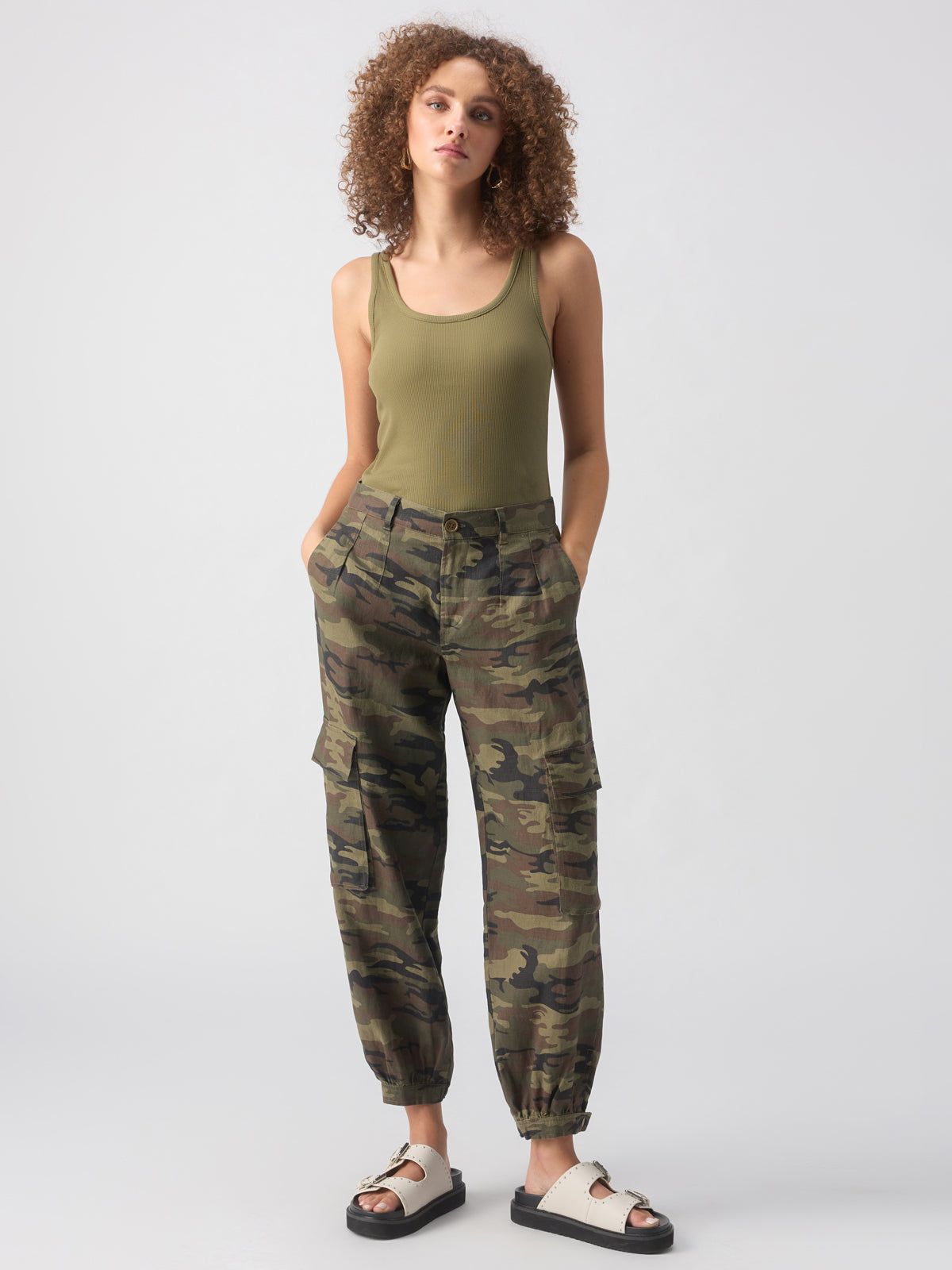 A person with curly hair wears a sleeveless olive green tank top and the Pleated Linen Cargo Little Hero Camo pants by Sanctuary Clothing. They stand with hands in pockets and feet slightly apart in a relaxed pose, also wearing white slide sandals with black soles. The background is plain white.