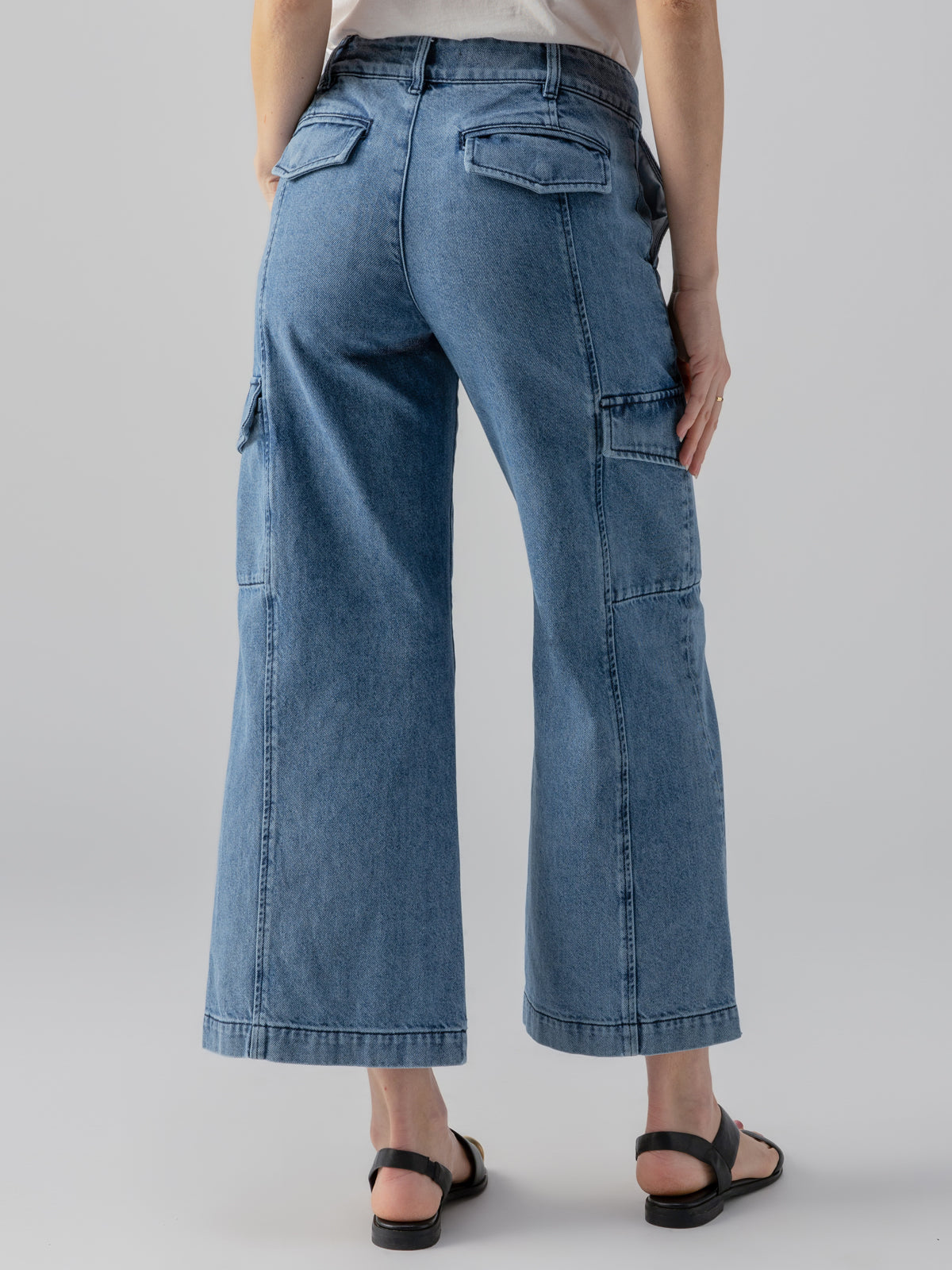 Back view of a person wearing the Rebel Wide Leg Crop Pant in Hazy Blue by Sanctuary Clothing, paired with a white shirt. The person is also wearing black sandals and standing against a plain gray background.