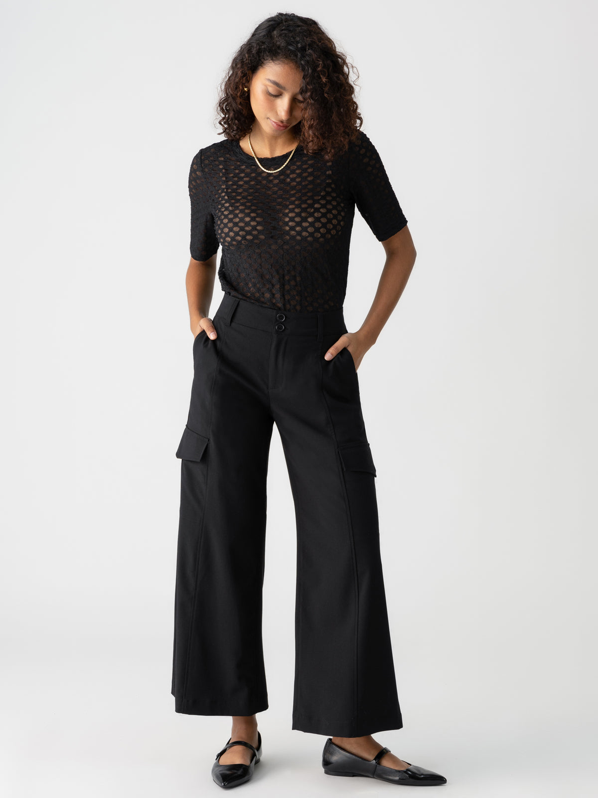 A person with curly hair is looking down while posing against a white background. They are wearing a black, see-through, short-sleeved top, Sanctuary Clothing's "Rebel Wide Leg Crop Black" high-waisted wide-leg pants with side pockets, and black flat shoes. Their hands are in their pockets.