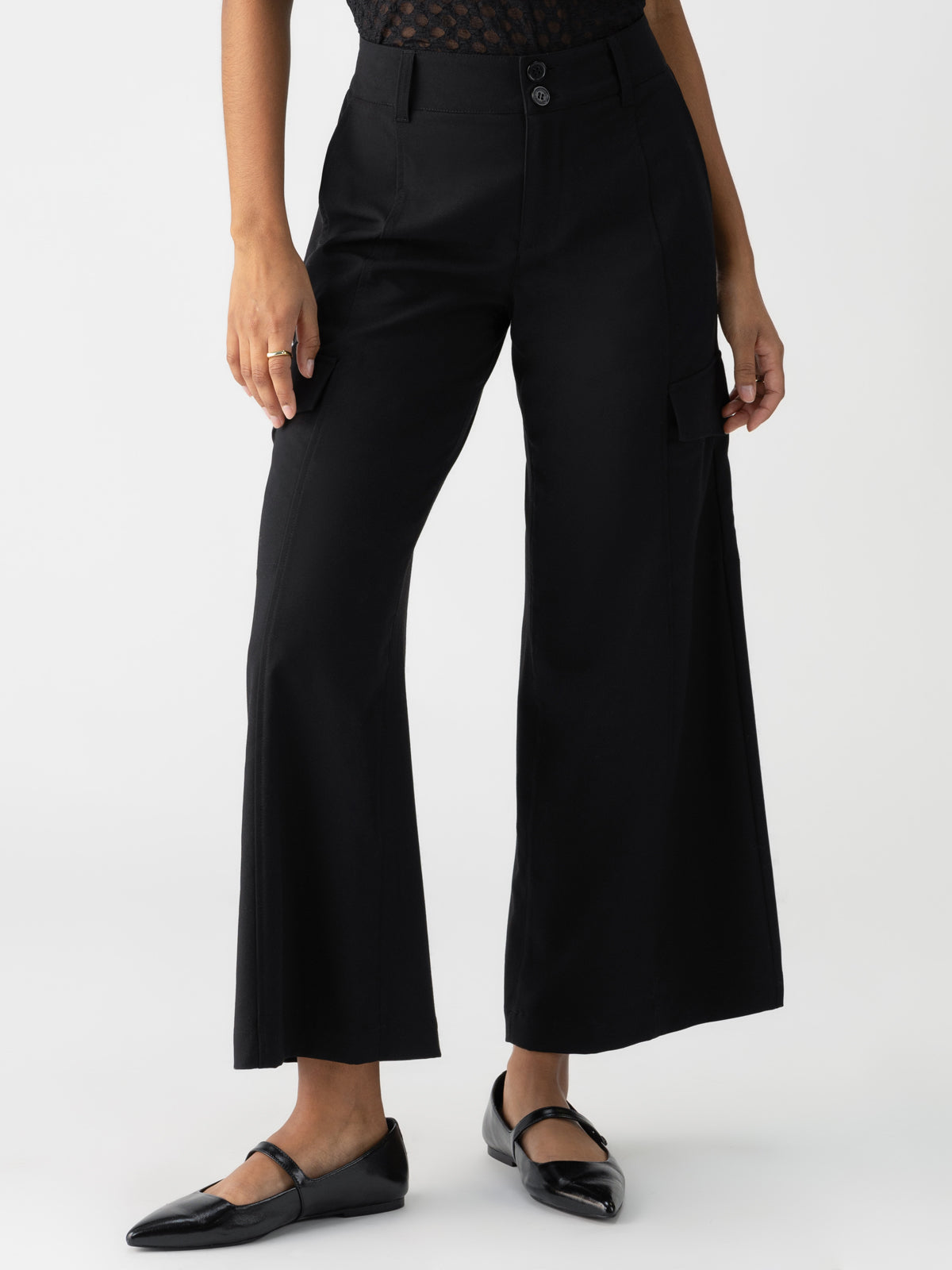 A person is wearing the Rebel Wide Leg Crop Black pants from Sanctuary Clothing. The high-waisted, wide-leg pants feature a double-button closure and a cargo pocket on one side. They are paired with a sheer patterned black top and black pointed-toe flats. The person stands with one hand by their side.