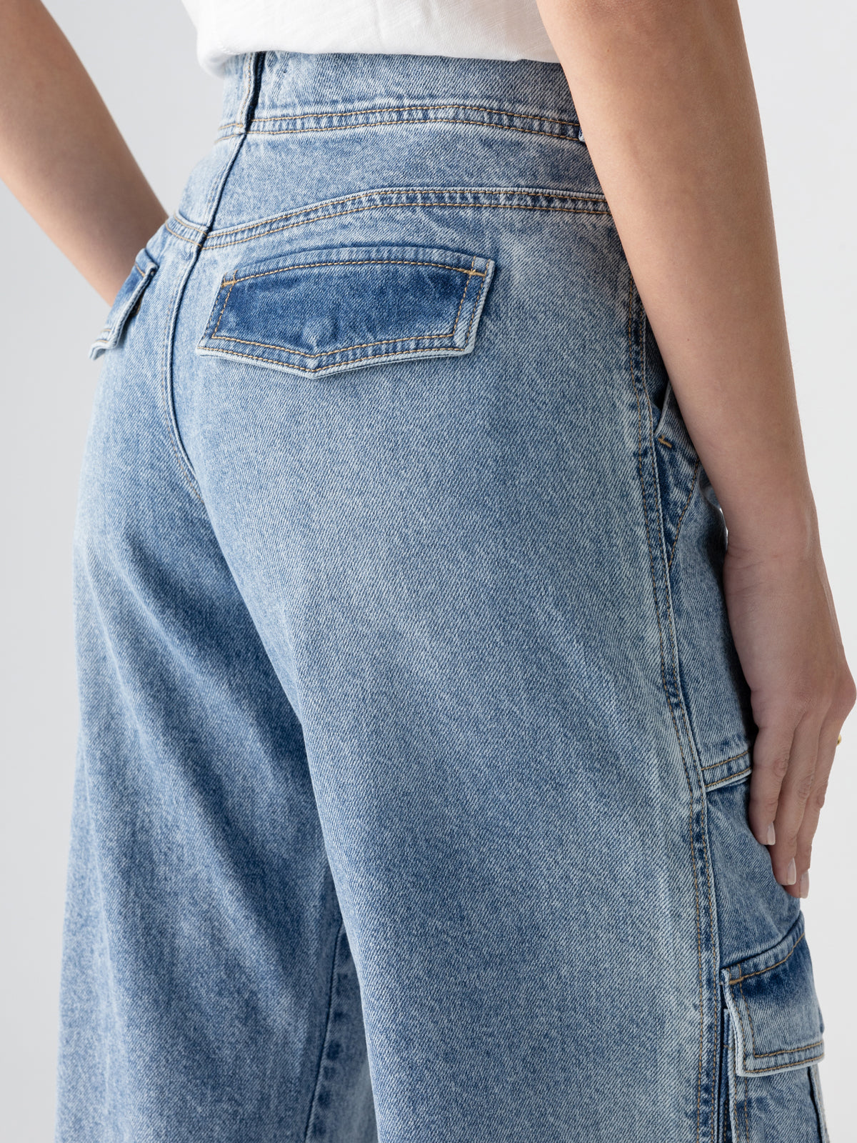 A close-up of a person wearing Sanctuary Clothing's Level Up Denim Cargo Pant in Digital Blue, featuring a back pocket and side cargo pocket. The person is standing sideways with their hand resting near their hip, partially showing a white top. These jeans have a relaxed, casual fit.