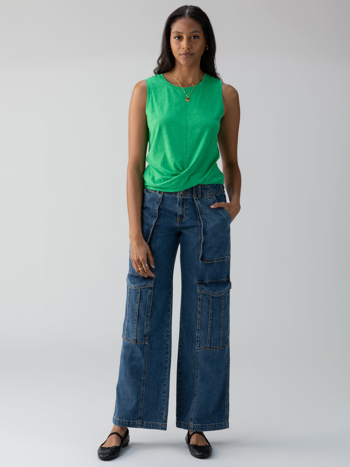 A woman stands against a neutral background wearing a sleeveless green top and Sanctuary Clothing's Level Up Cargo Pant Oasis, featuring wide-legged denim and multiple pockets. She has one hand in her pocket and wears black flats, with a necklace featuring a small pendant. She has a neutral expression.