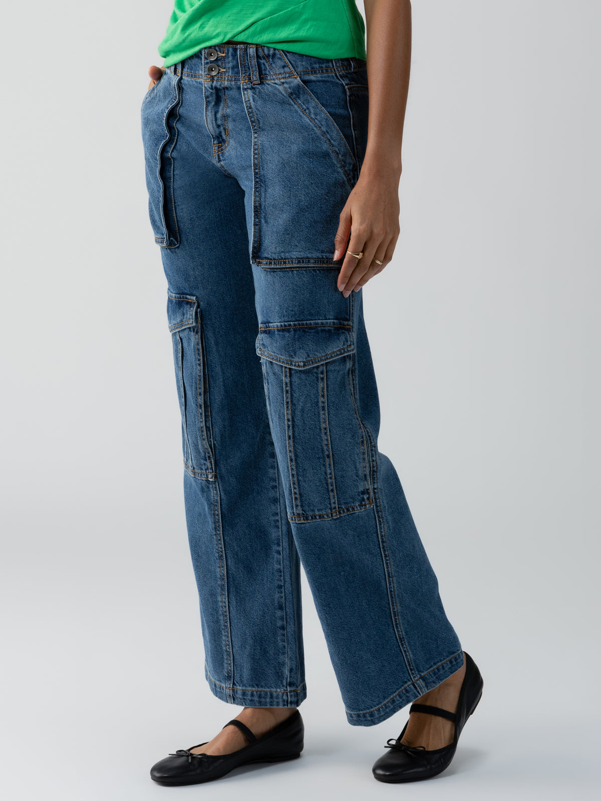 A person wearing the Sanctuary Clothing Level Up Cargo Pant Oasis, which are blue denim cargo pants with multiple pockets, paired with a green top. They also have on black ballet flats. The background is plain and light-colored.