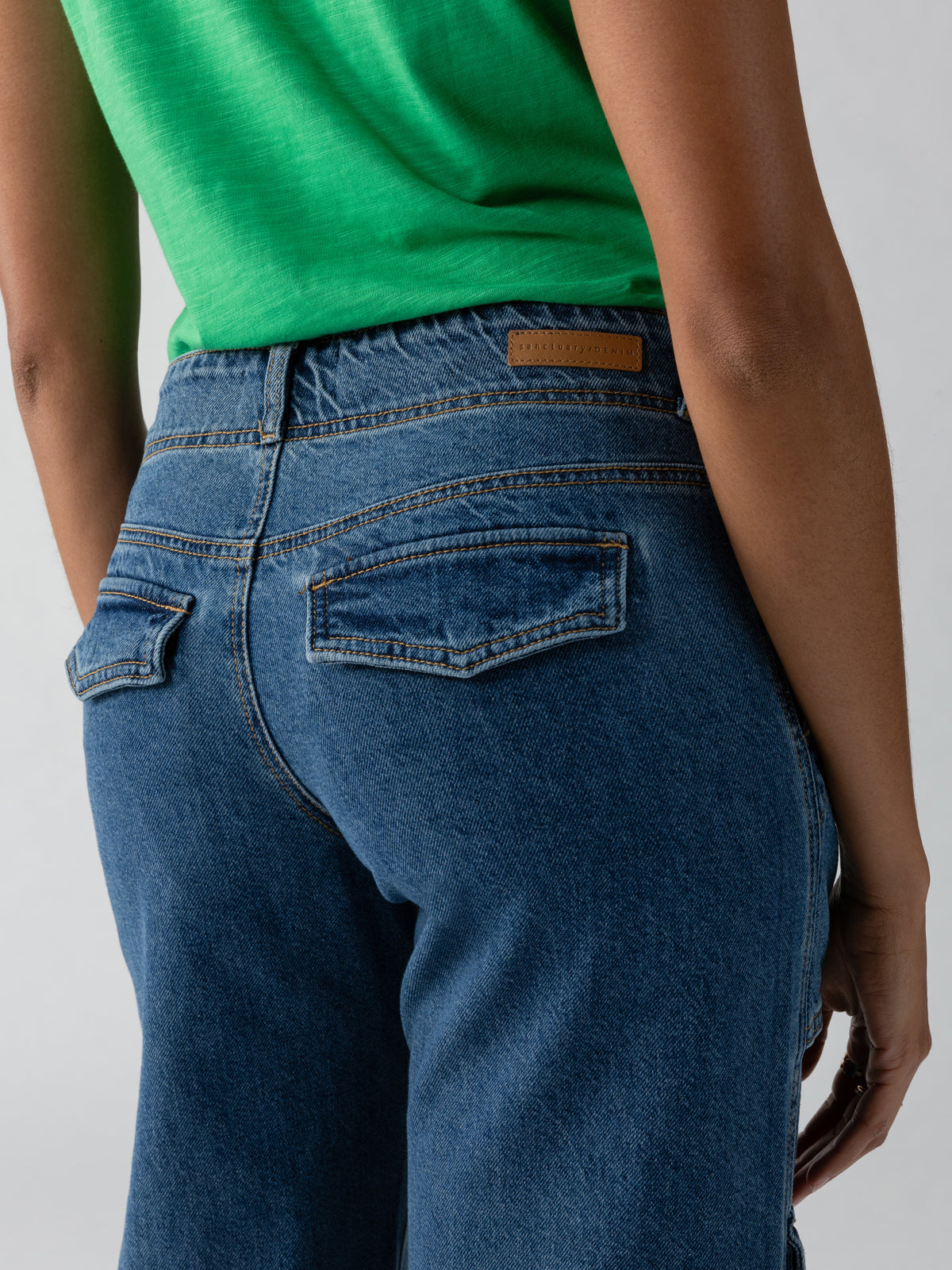 A person wearing Level Up Cargo Pant Oasis from Sanctuary Clothing and a green top, photographed from the back. The cargo pants have two back pockets with flaps and stitching details. The background is plain and light-colored.