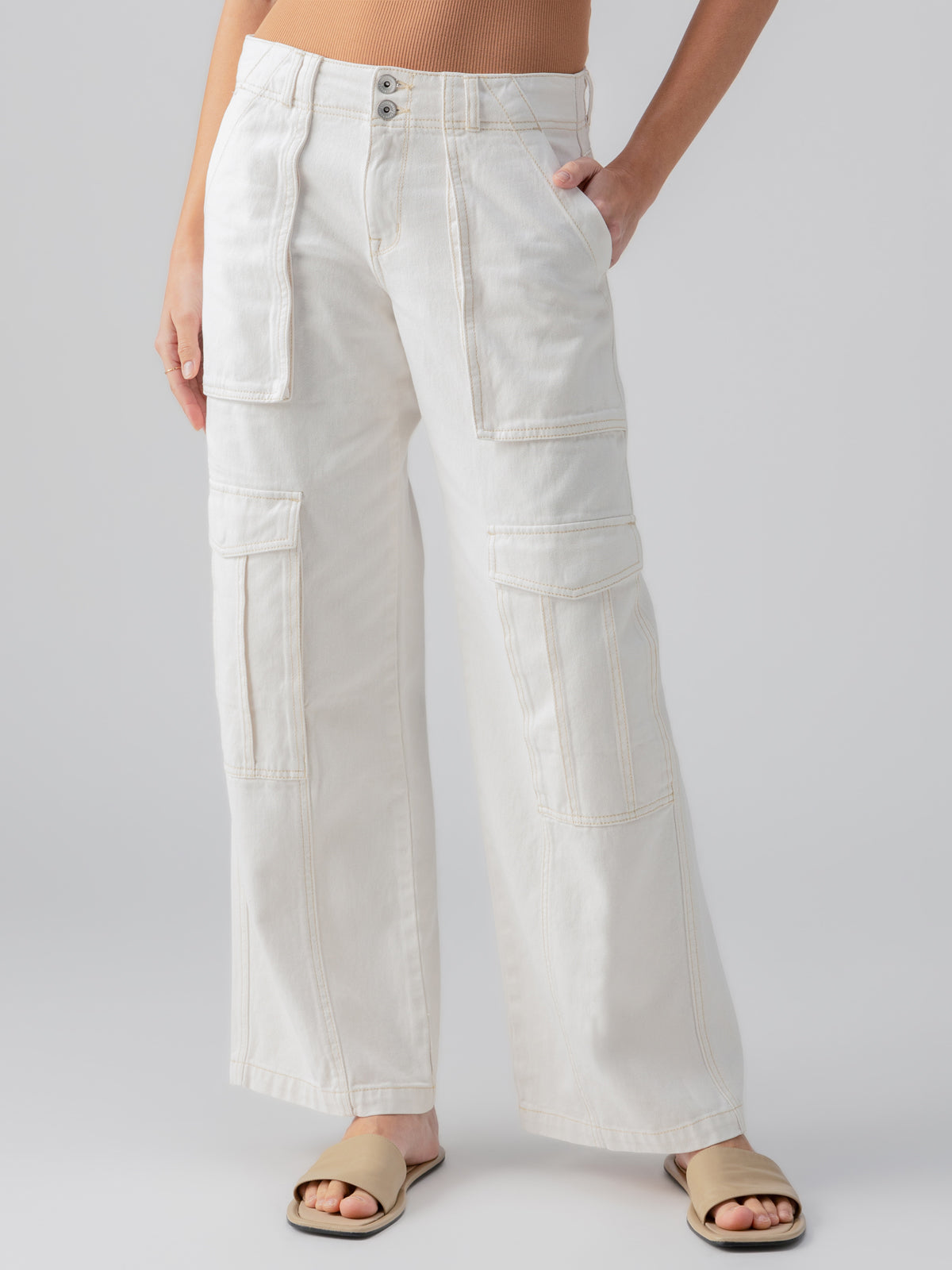 A person is modeling Sanctuary Clothing's Level Up Cargo Pant in Powdered Sugar, featuring a high-waisted, wide-leg design with multiple large pockets. They pair it with a beige cropped top and beige slip-on sandals. The person's right hand rests in a pocket while their left arm is relaxed by their side.