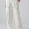 A person is modeling Sanctuary Clothing's Level Up Cargo Pant in Powdered Sugar, featuring a high-waisted, wide-leg design with multiple large pockets. They pair it with a beige cropped top and beige slip-on sandals. The person's right hand rests in a pocket while their left arm is relaxed by their side.