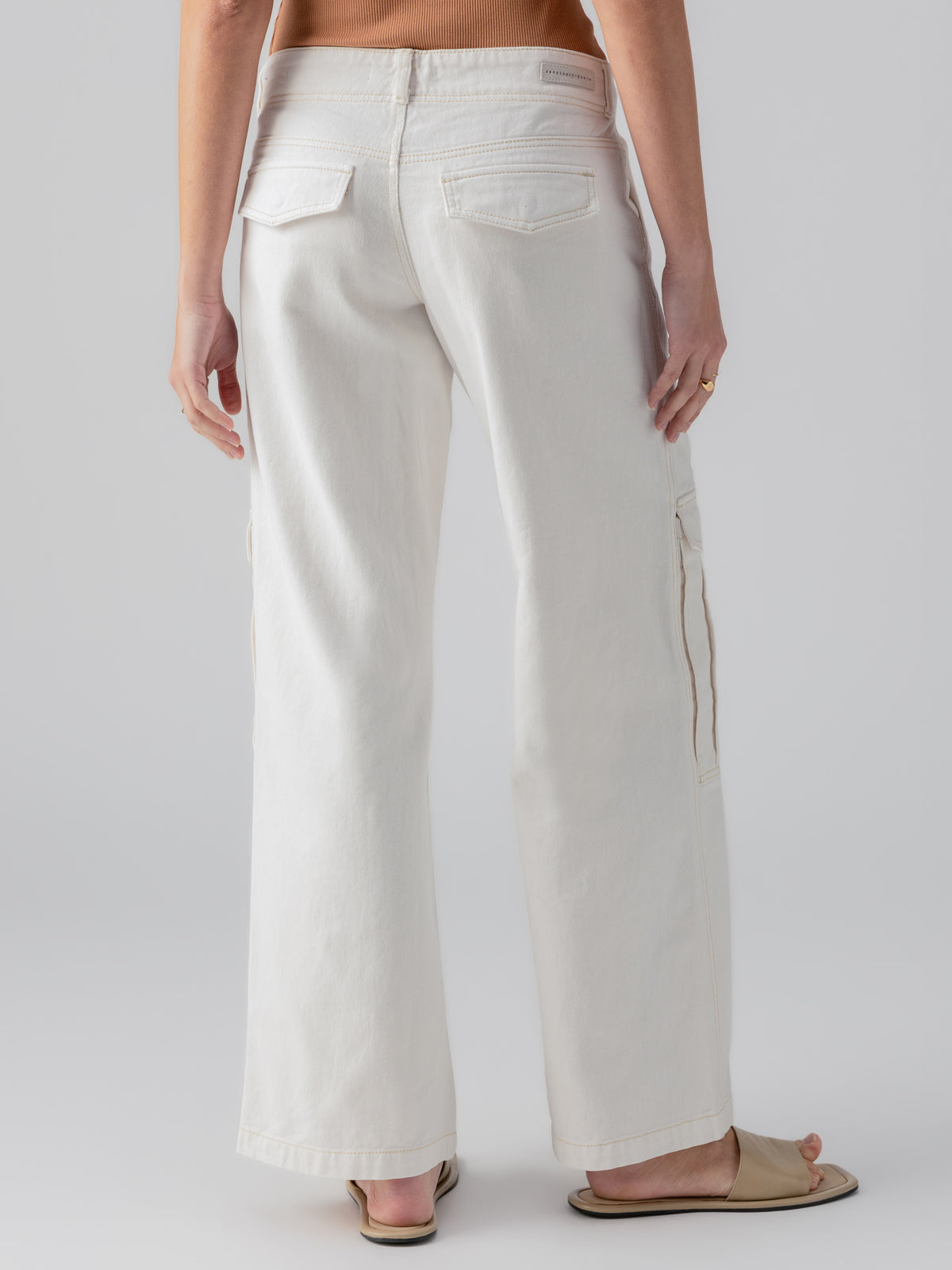 A person is shown from the back wearing the Level Up Cargo Pant in Powdered Sugar by Sanctuary Clothing. The pants are high-waisted with wide legs and feature side and back pockets. They are also wearing beige sandals and have one hand resting by their side. The background is plain and light gray.