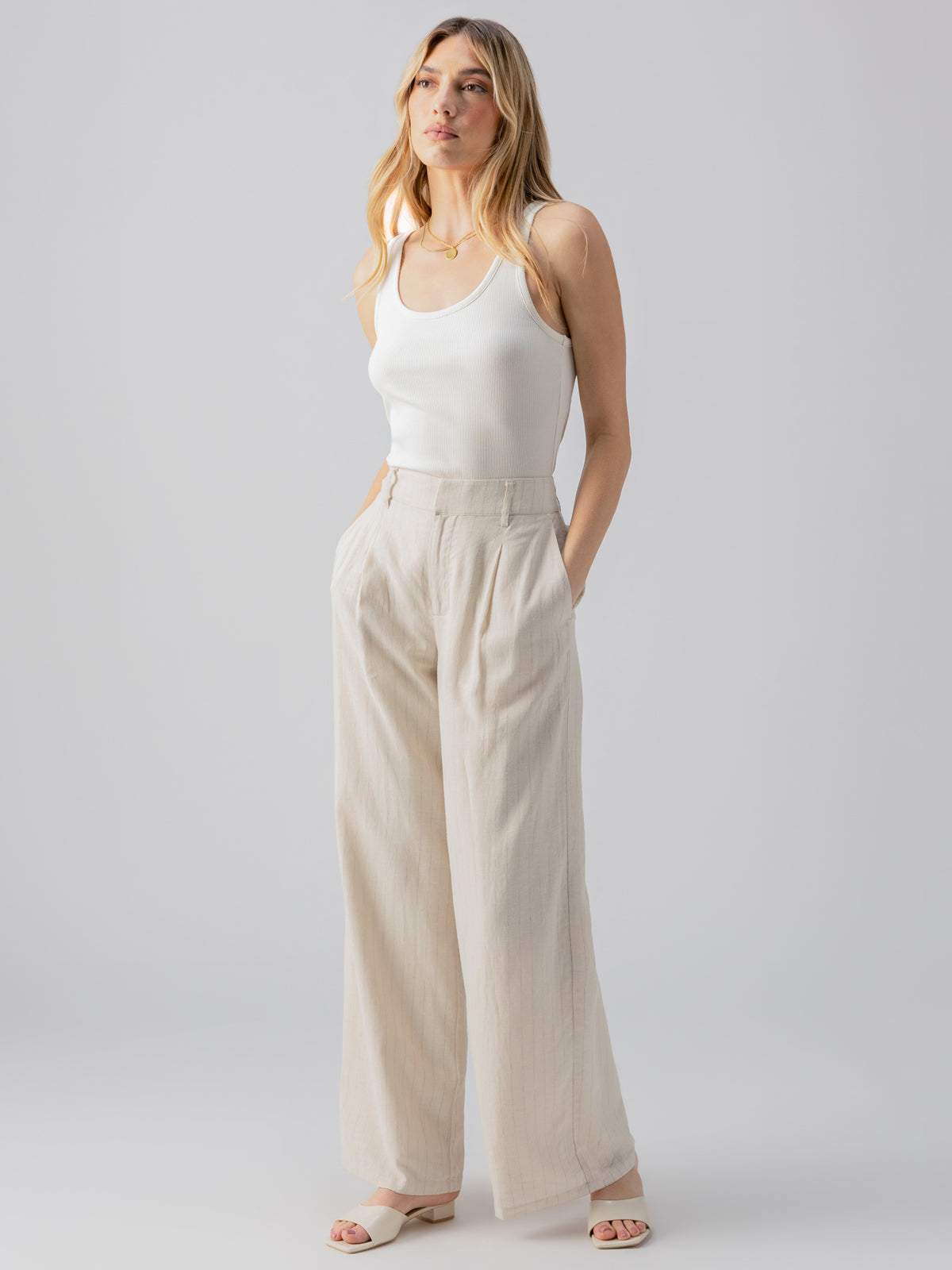 A person with light hair is standing against a light grey background. They are wearing a white sleeveless top and the Pleat Up Trouser Vineyard Stripe by Sanctuary Clothing. Their hands are in their pockets, and they are looking slightly off to the side.