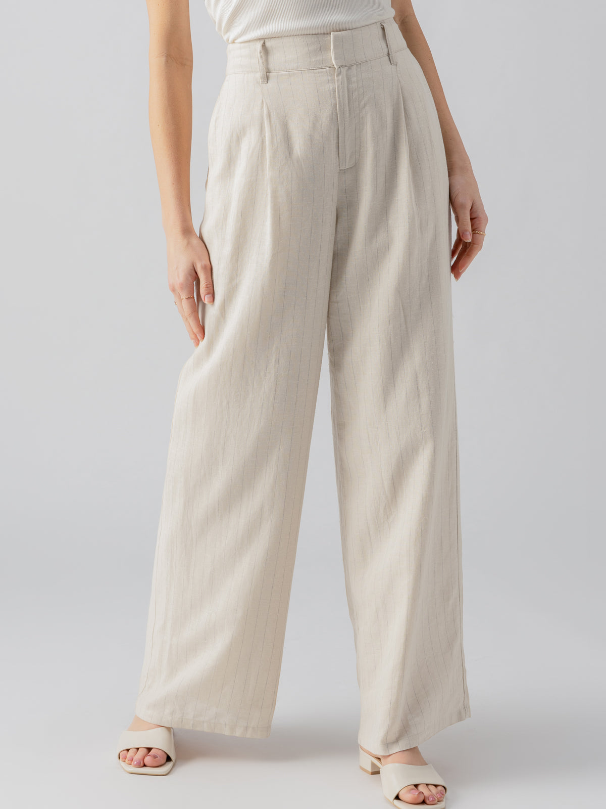 A person is wearing Sanctuary Clothing's Pleat Up Trouser Vineyard Stripe, featuring light beige wide-leg pants with a high waist and vertical pleats. The person is also wearing a sleeveless white top and open-toed, low-heeled white sandals. The background is plain and greyish.