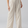 A person is wearing Sanctuary Clothing's Pleat Up Trouser Vineyard Stripe, featuring light beige wide-leg pants with a high waist and vertical pleats. The person is also wearing a sleeveless white top and open-toed, low-heeled white sandals. The background is plain and greyish.