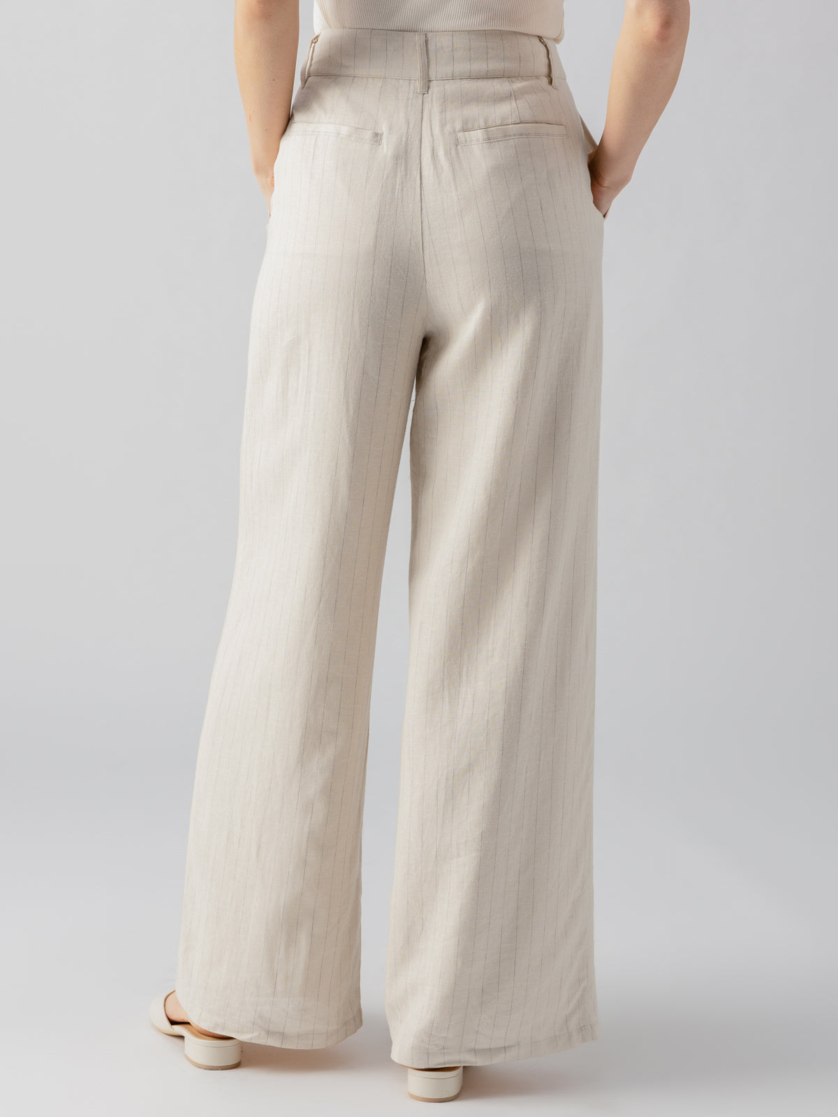 A person is standing with their back to the camera, wearing the Pleat Up Trouser Vineyard Stripe from Sanctuary Clothing. The beige trousers feature subtle vertical stripes and a wide-leg cut that nearly touches the ground. They are paired with light-colored shoes, set against a plain, light grey background.