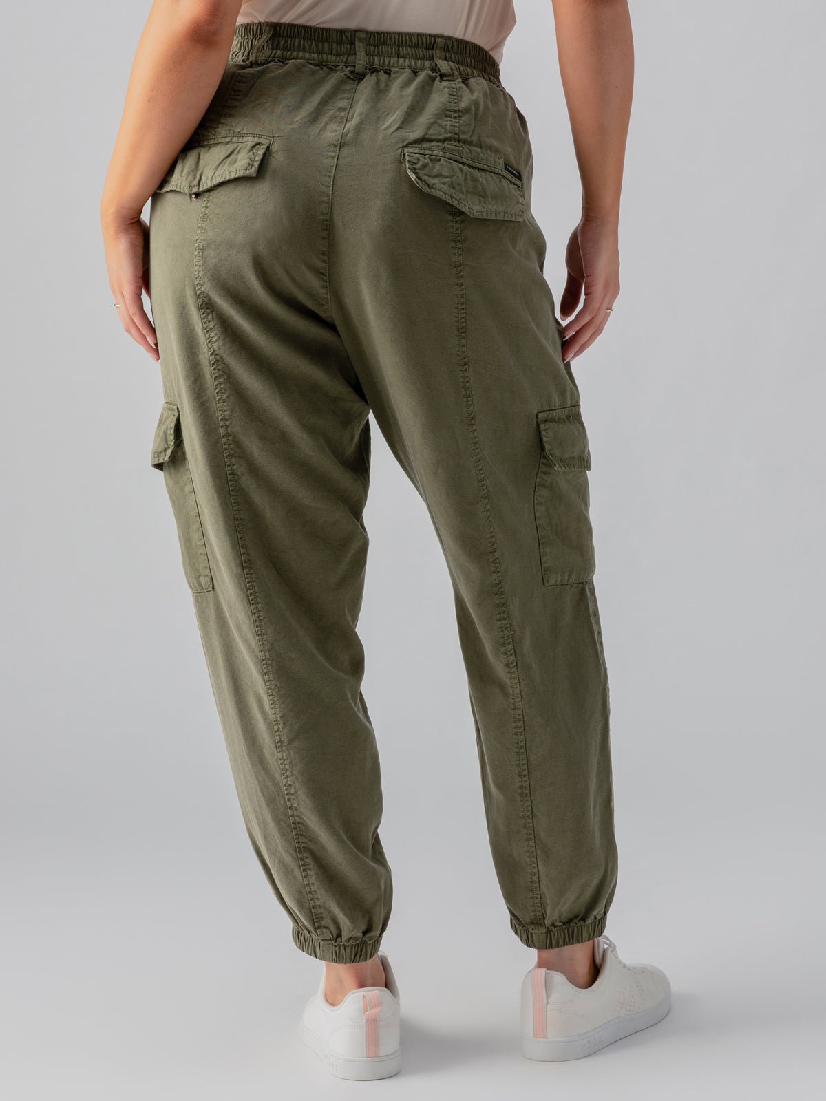 A person is standing with their back to the camera, wearing the Relaxed Rebel Burnt Olive cargo pants from Sanctuary Clothing's Inclusive Collection. The pants feature multiple pockets and elastic cuffs, paired with white sneakers accented in pink. The background is plain and gray.