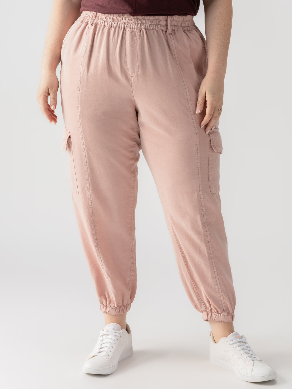 A person is seen from the waist down, wearing the "Relaxed Rebel Smokey Rose" cargo jogger pants from Sanctuary Clothing's Inclusive Collection. The light pink joggers feature side pockets and elastic cuffs at the ankles, paired with white sneakers against a plain, light-colored background.