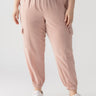 A person is seen from the waist down, wearing the "Relaxed Rebel Smokey Rose" cargo jogger pants from Sanctuary Clothing's Inclusive Collection. The light pink joggers feature side pockets and elastic cuffs at the ankles, paired with white sneakers against a plain, light-colored background.