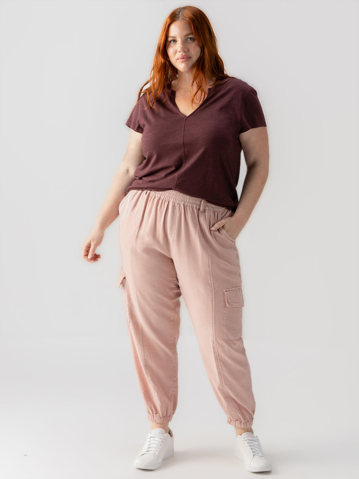 A woman with long reddish hair stands against a plain background, showcasing the "Relaxed Rebel Smokey Rose Inclusive Collection" from Sanctuary Clothing. She is wearing a maroon V-neck T-shirt paired with light pink jogger pants that feature convenient side pockets and white sneakers. Her hands are comfortably in her pockets, exuding a relaxed and confident demeanor.