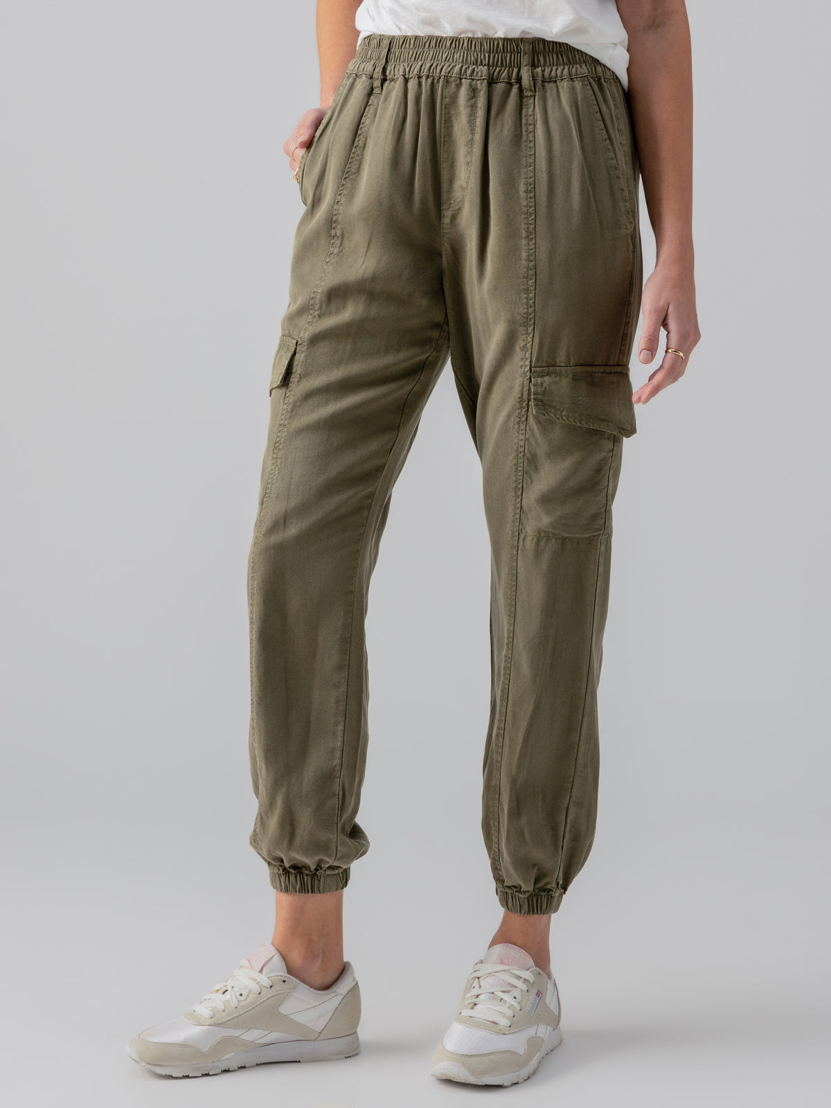 A person is wearing Sanctuary Clothing's Relaxed Rebel Burnt Olive cargo pants with elastic cuffs and side pockets. They are also wearing light-colored sneakers and a white top. Only the lower half of the person’s body is visible against a plain, light-colored background.