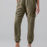 A person is wearing Sanctuary Clothing's Relaxed Rebel Burnt Olive cargo pants with elastic cuffs and side pockets. They are also wearing light-colored sneakers and a white top. Only the lower half of the person’s body is visible against a plain, light-colored background.