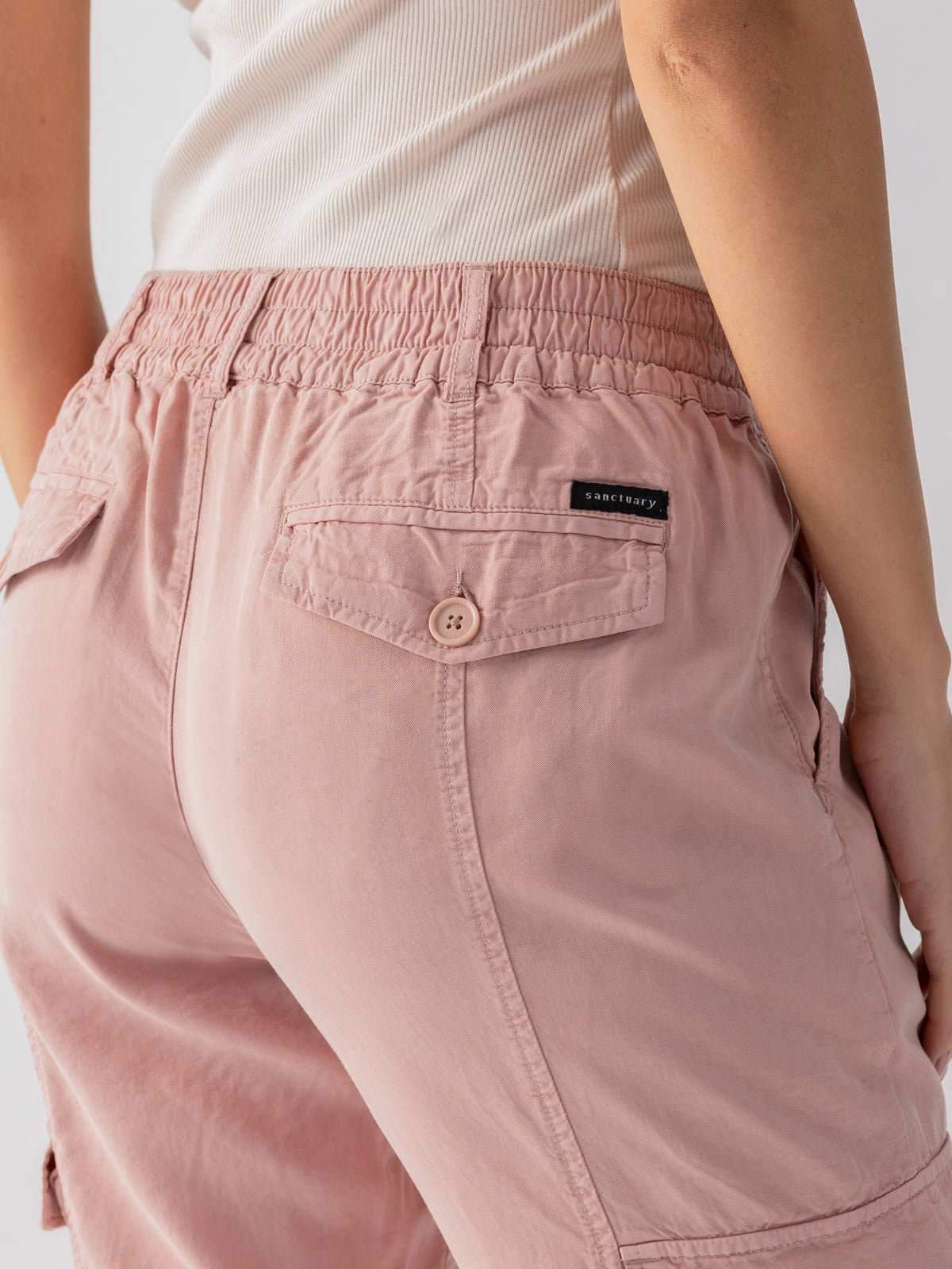 A person is shown from the waist down wearing Relaxed Rebel Smokey Rose cargo pants from Sanctuary Clothing, featuring an elastic waistband and a back pocket with a button closure and a small black label. They pair the light pink pants with a white sleeveless top.