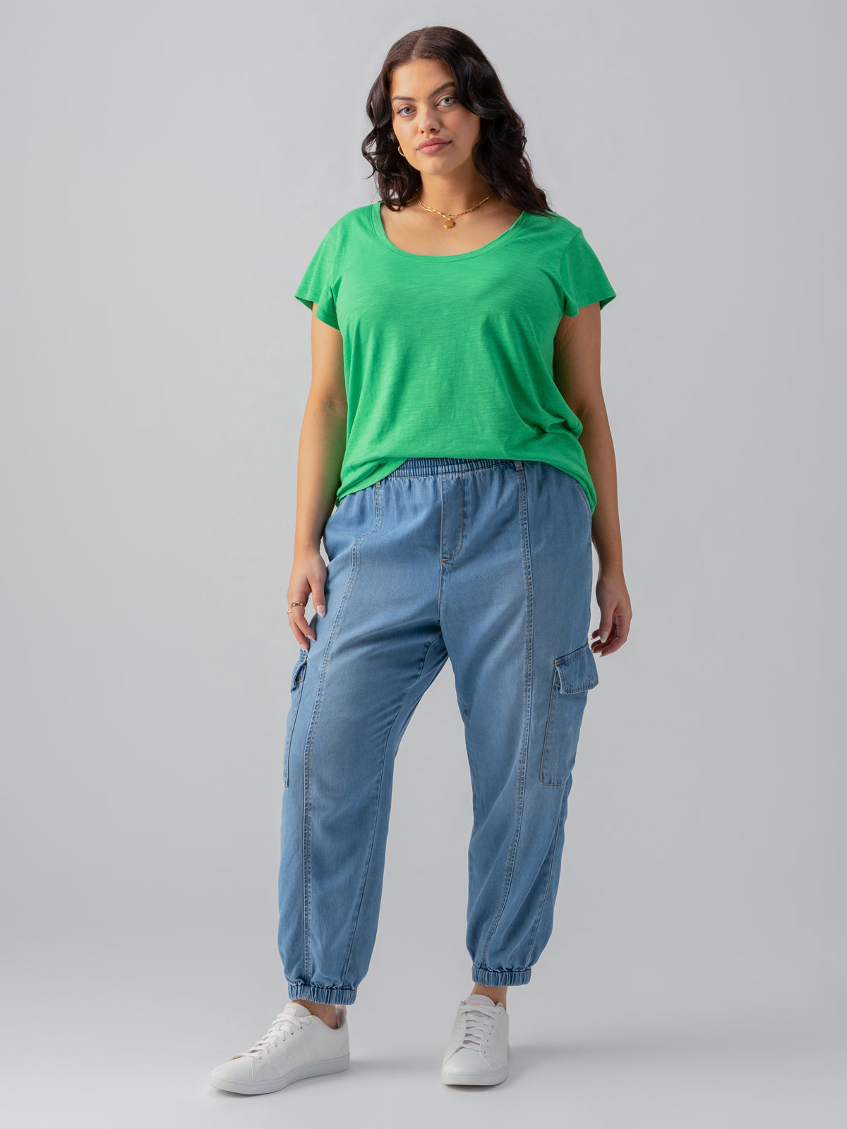 A person with long wavy hair is standing against a plain background, wearing a green short-sleeve top and light blue cargo jogger pants from the Relaxed Rebel Sun Drenched Inclusive Collection by Sanctuary Clothing. They have white sneakers on and their hands are casually positioned in their pockets. They also have a neutral expression.