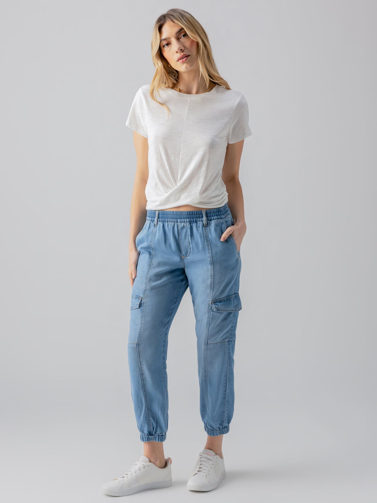 A woman stands against a plain grey background, wearing the Relaxed Rebel Sun Drenched t-shirt by Sanctuary Clothing, tucked into light blue jogger-style cargo pants. She has one hand in her pocket and is looking directly at the camera, completing her look with white sneakers.