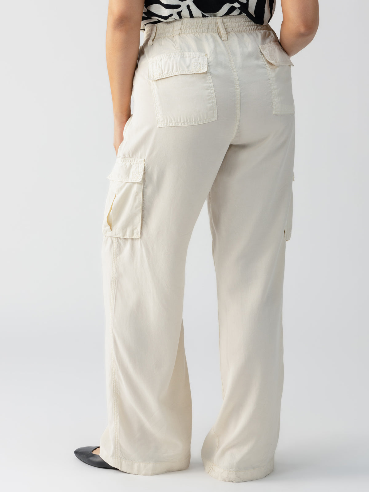 A person is shown from the back, wearing Relaxed Reissue Birch Inclusive Collection cargo pants by Sanctuary Clothing. The pants are light-colored with multiple pockets, paired with a black and white patterned top. The person stands slightly leaning forward, their left hand resting on their hip, and they wear black shoes.