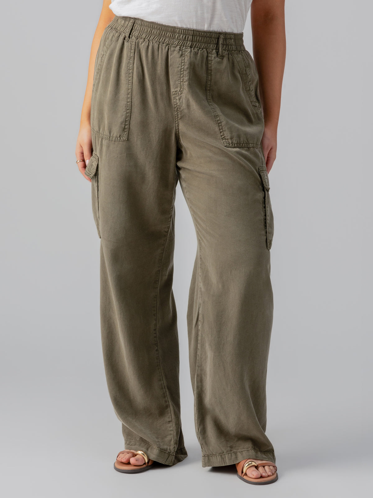 A person is wearing the Relaxed Reissue Burnt Olive Inclusive Collection pants from Sanctuary Clothing, which feature an elastic waist and large front pockets, paired with a white top. They are also sporting tan sandals against a plain gray background.