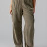 A person is wearing the Relaxed Reissue Burnt Olive Inclusive Collection pants from Sanctuary Clothing, which feature an elastic waist and large front pockets, paired with a white top. They are also sporting tan sandals against a plain gray background.