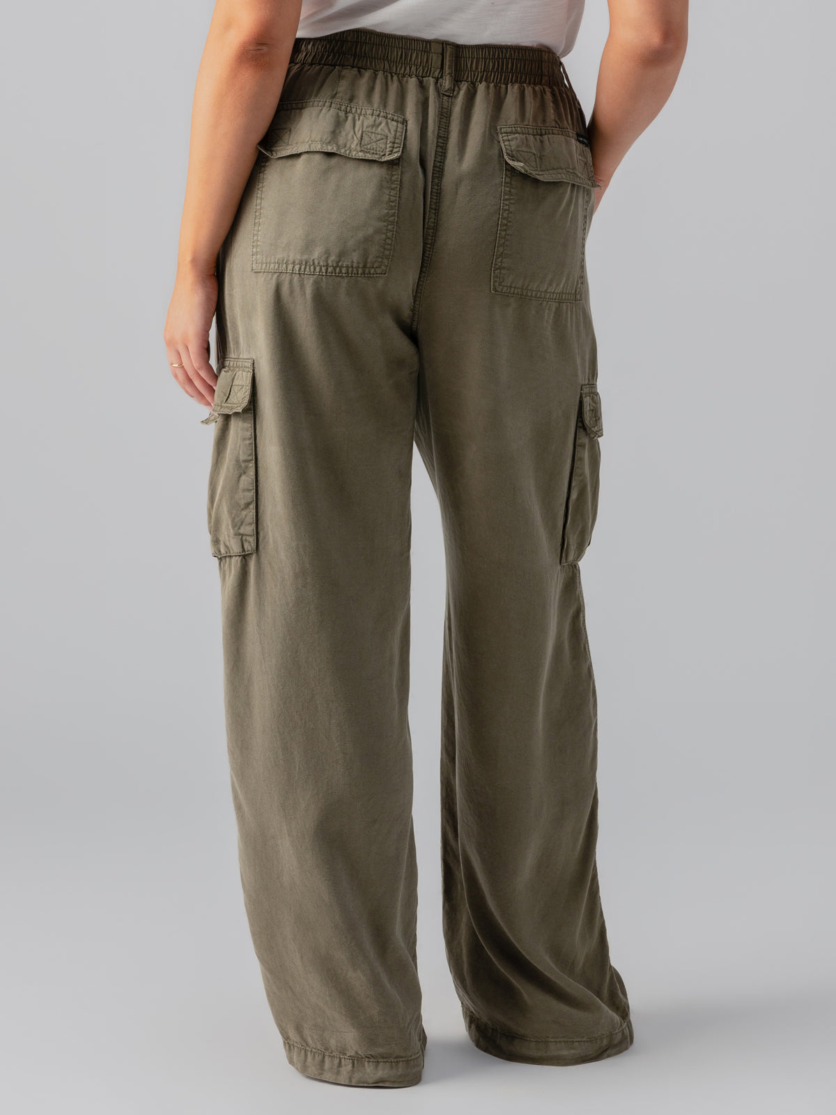 A person is seen from the back, wearing the "Relaxed Reissue Burnt Olive" wide-leg cargo pants from Sanctuary Clothing's Inclusive Collection, featuring large pockets on the sides and back. The waistband is elastic, and the pants have a relaxed fit. The person is also wearing a light-colored top.