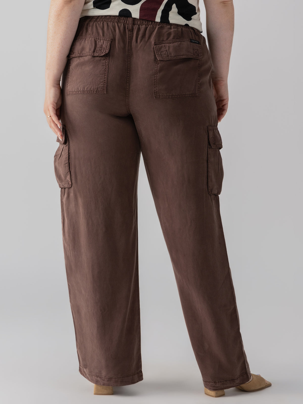 A person is standing with their back to the camera, showcasing items from the Sanctuary Clothing Relaxed Reissue Mud Bath Inclusive Collection. They are wearing brown cargo pants featuring flap pockets on the back and sides, paired with a beige and dark red patterned top. Beige shoes peek out from beneath the pants. The background is plain and light-colored.