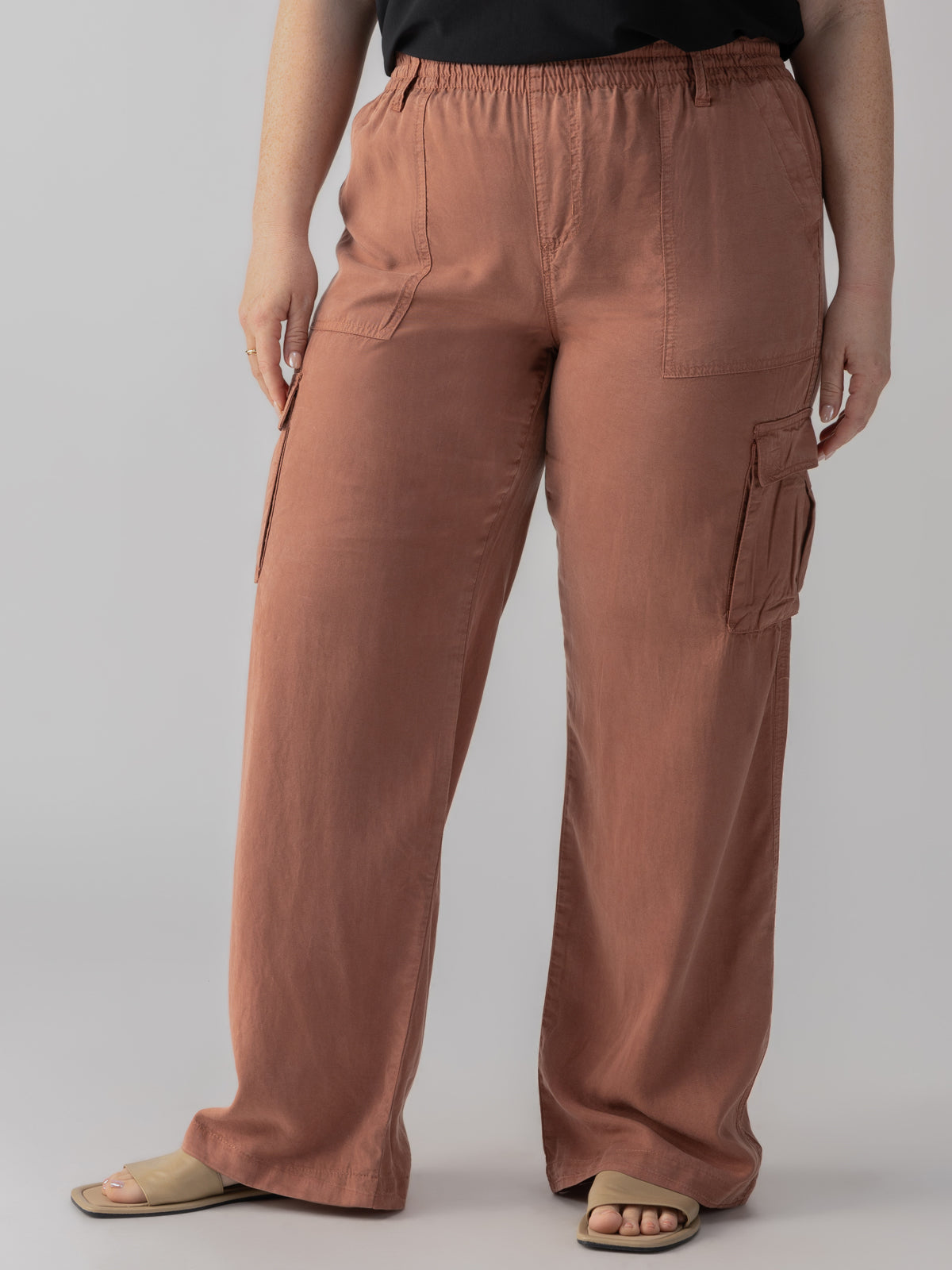 A person is wearing the Relaxed Reissue Washed Clay Inclusive Collection pants by Sanctuary Clothing. These loose-fitting, high-waisted cargo pants are rust-colored and made of lightweight fabric. They feature large side pockets and straight legs. The person is also wearing a black top and light-colored slide sandals, with only the lower half of their body visible.