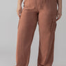 A person is wearing the Relaxed Reissue Washed Clay Inclusive Collection pants by Sanctuary Clothing. These loose-fitting, high-waisted cargo pants are rust-colored and made of lightweight fabric. They feature large side pockets and straight legs. The person is also wearing a black top and light-colored slide sandals, with only the lower half of their body visible.